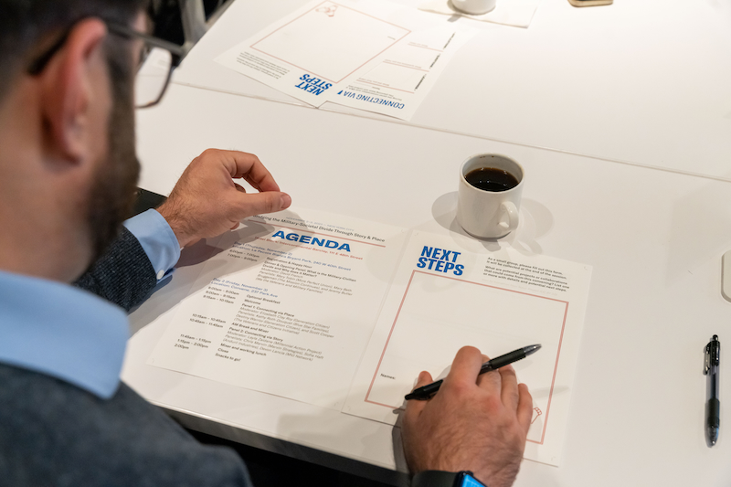 A person with glasses is sitting at a table, holding a pen and looking at an agenda and a 'Next Steps' worksheet. A cup of coffee is on the table.