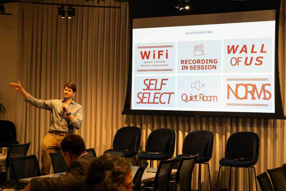 Person speaking into a microphone and gesturing towards a presentation screen displaying various housekeeping notes.