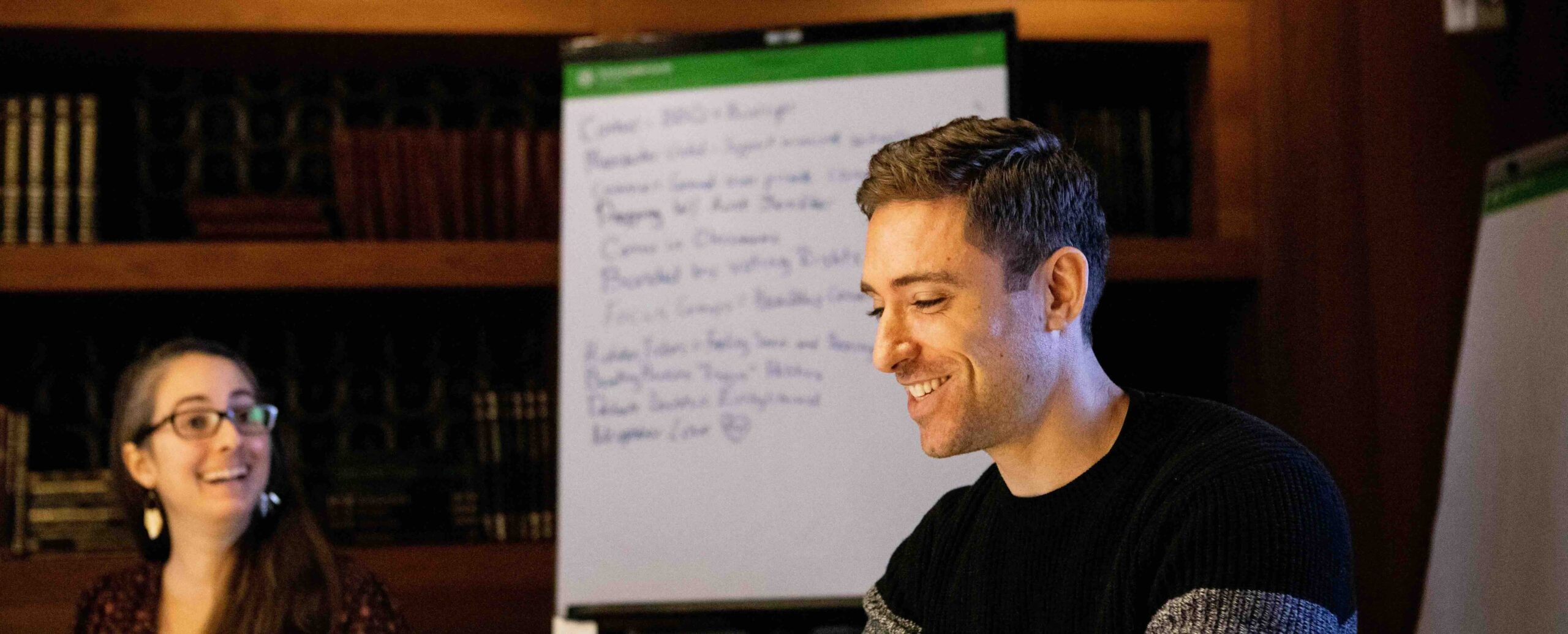 Two people are smiling and talking at a table in a library, with a flipchart in the background.