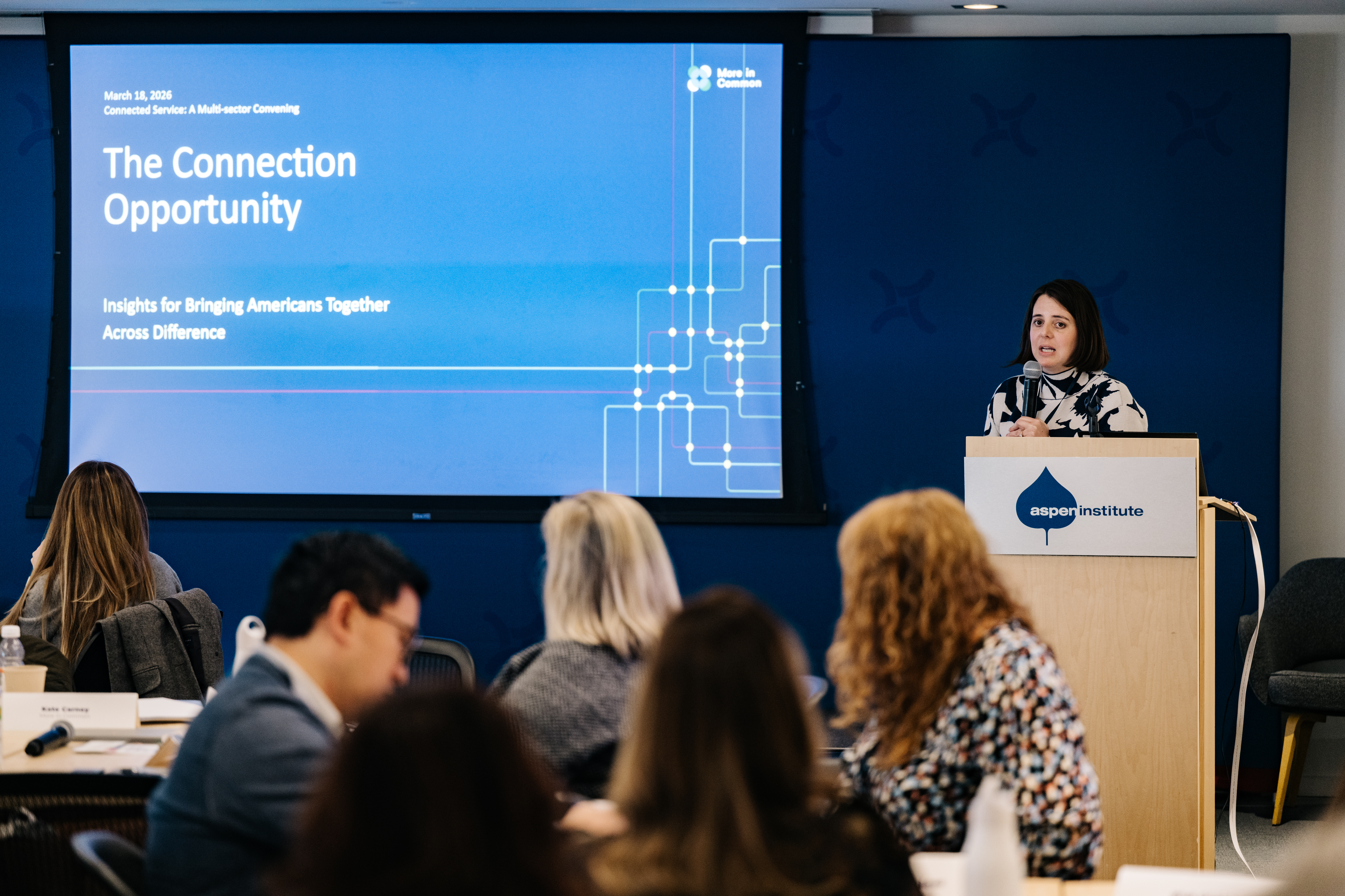 Woman speaking into a microphone at a podium next to a presentation screen about The Connection Opportunity report. 